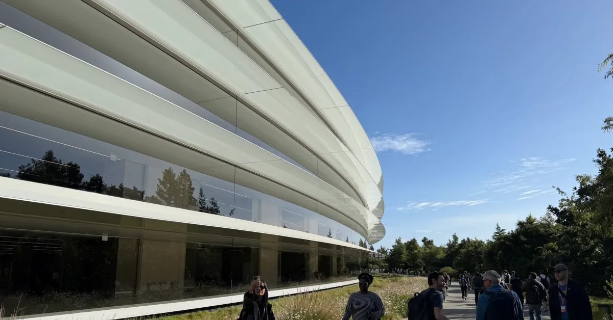 Apple Park i Cupertino: den svängda glasfasaden speglar träden medan besökare går längs gången utanför en solig dag.