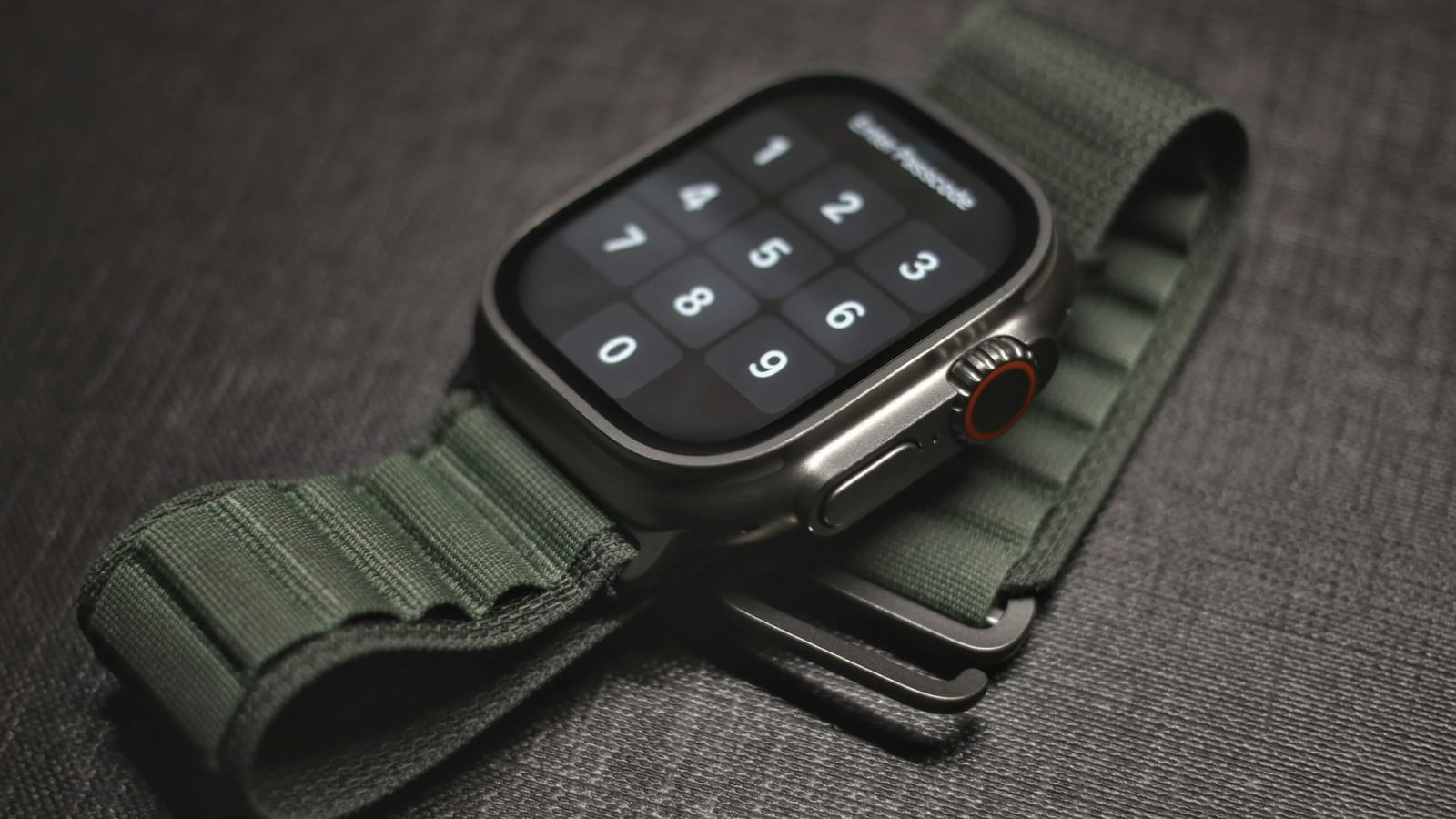 Apple Watch Ultra i titan med grön Alpine Loop-rem, liggande på mörkt tyg med kodlåsskärmen tänd.