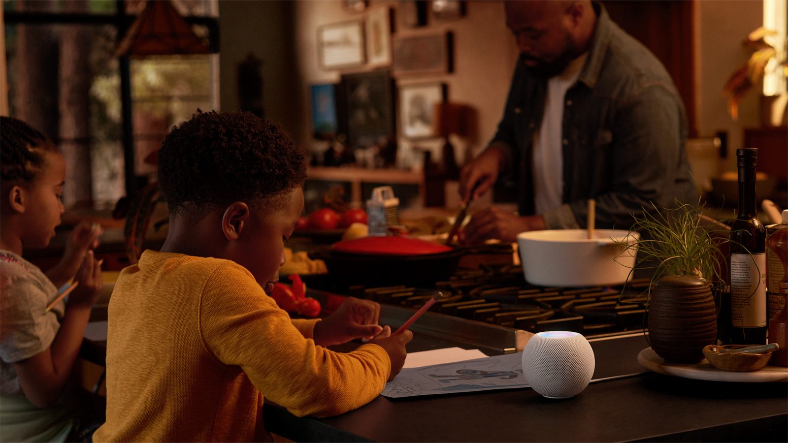 En vit HomePod mini står på en köksbänk medan ett barn ritar och en vuxen lagar mat i bakgrunden.