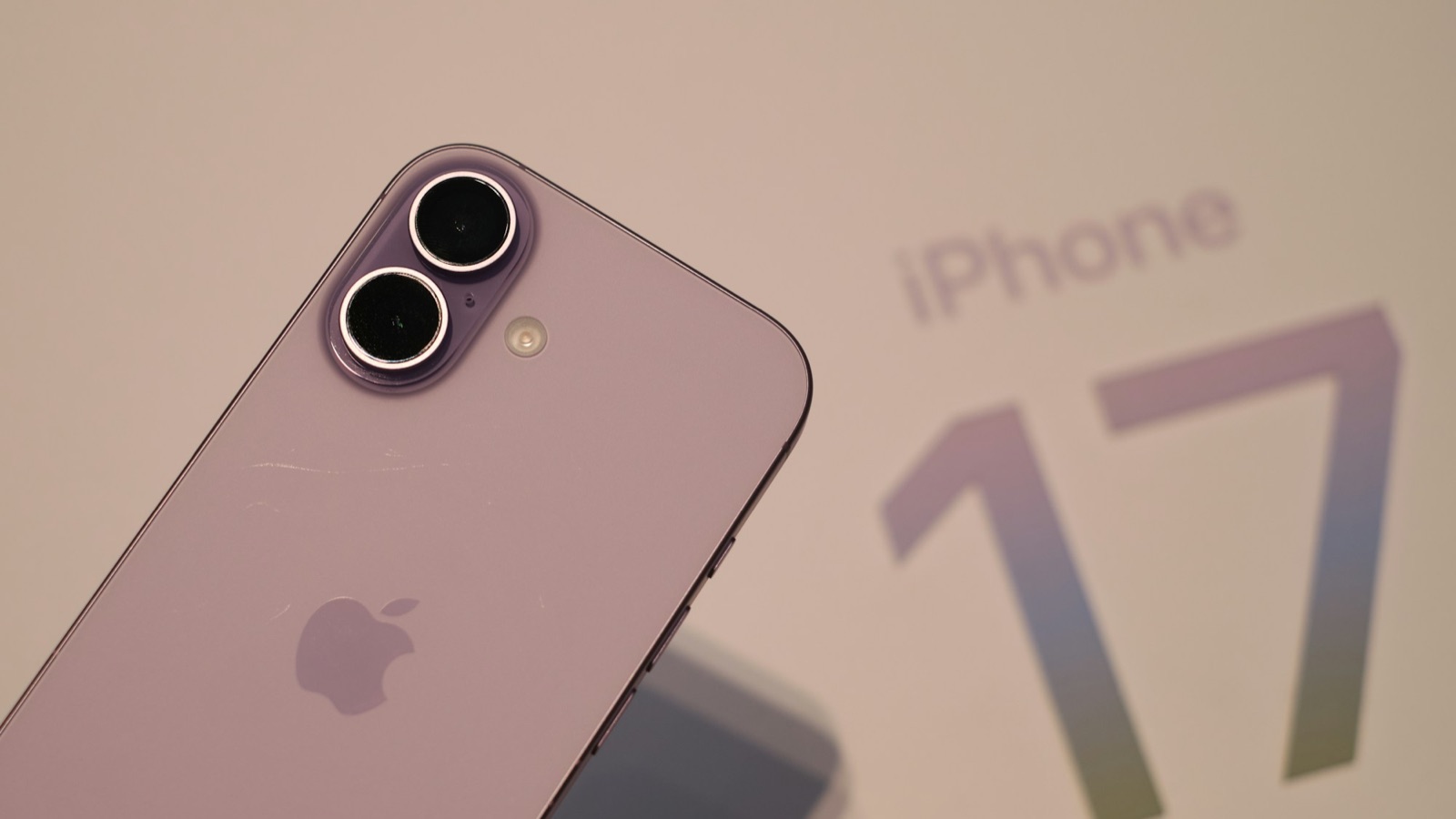 Baksidan av en lavendelfärgad iPhone 17 med dubbla kameror framför en iPhone 17-skylt